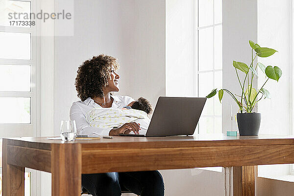 Frau mit Baby schaut durch das Fenster  während sie zu Hause im Büro sitzt