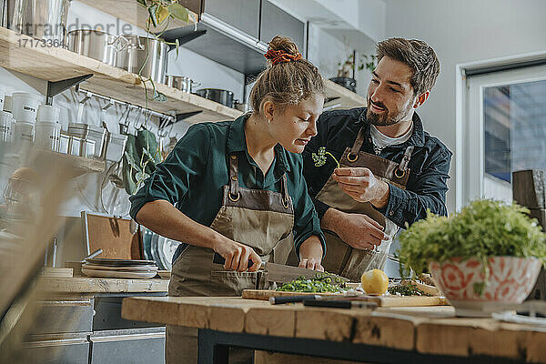 Young chef smelling parsley while cutting scallions standing by colleague on kitchen island
