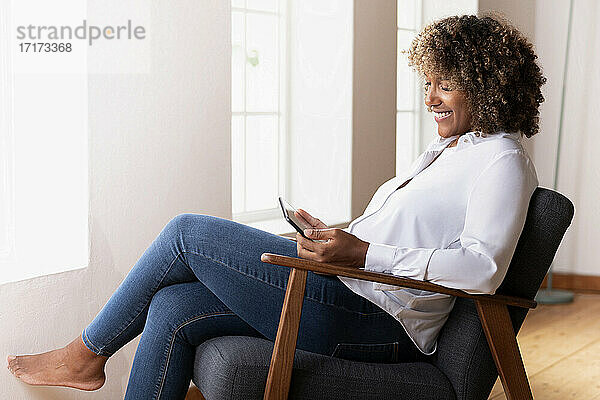 Lächelnde Frau  die ein Smartphone benutzt  während sie zu Hause auf einem Sessel sitzt