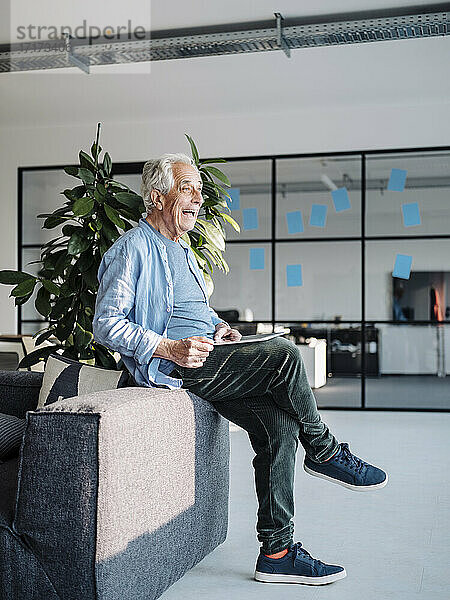 Älterer Geschäftsmann lächelt  während er im Büro sitzend ein digitales Tablet benutzt
