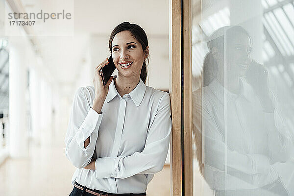 Lächelnde Geschäftsfrau  die mit ihrem Smartphone spricht  während sie sich im Büro an die Wand lehnt