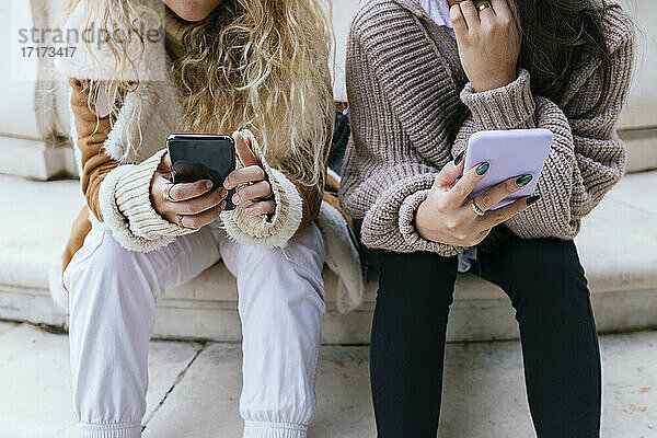 Female friends in warm clothing using mobile phone while sitting on steps