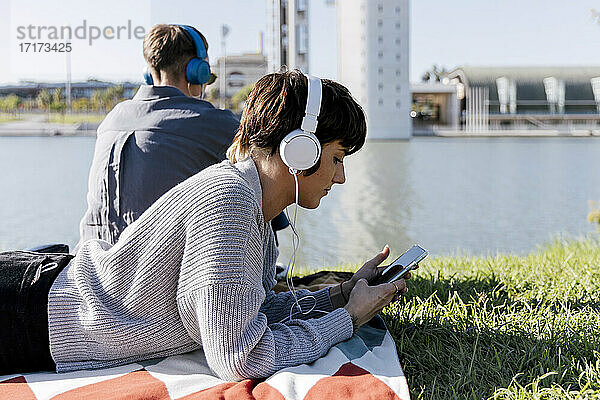 Frau mit Kopfhörern  die ein Mobiltelefon benutzt  während sie auf einer Decke neben einem Freund am Fluss liegt