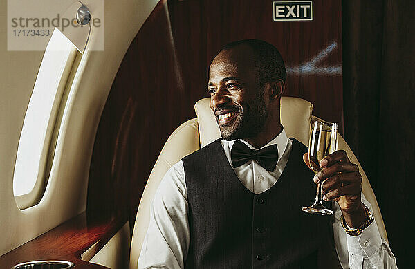 Lächelnder männlicher Unternehmer mit Champagner in der Hand  der aus dem Fenster eines Flugzeugs schaut
