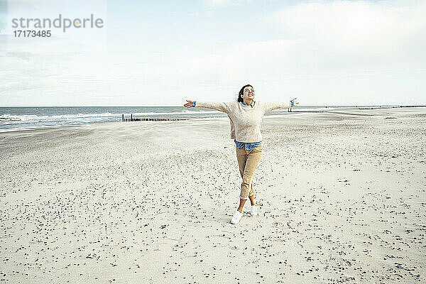 Porträt einer jungen Frau  die mit erhobenen Armen am Sandstrand spazieren geht