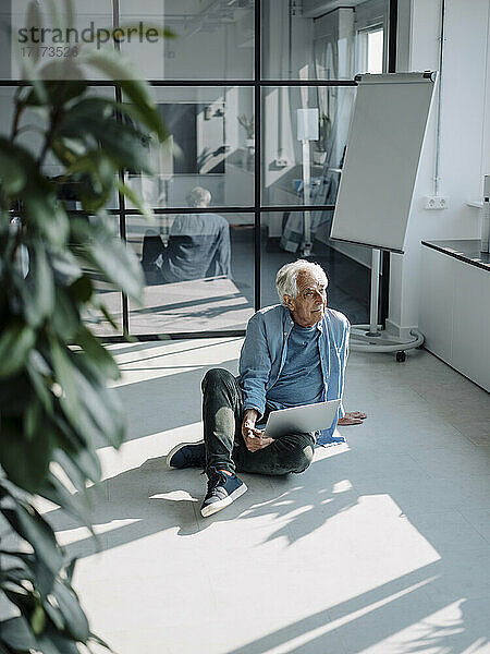 Senior Professional mit Laptop schaut weg  während er im Büro auf dem Boden sitzt