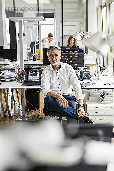 Älterer Geschäftsmann auf einem Stuhl sitzend mit einem im Hintergrund stehenden Kollegen in einem Großraumbüro