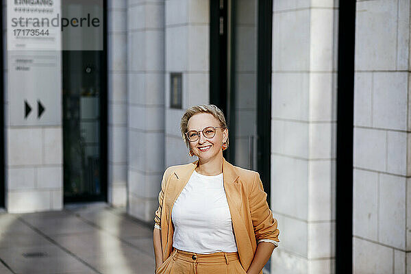 Lächelnde Geschäftsfrau mit Händen in den Taschen  die vor einem Bürogebäude steht