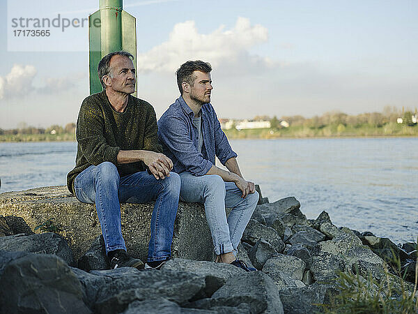 Thoughtful father and son looking away while sitting on rock against sky
