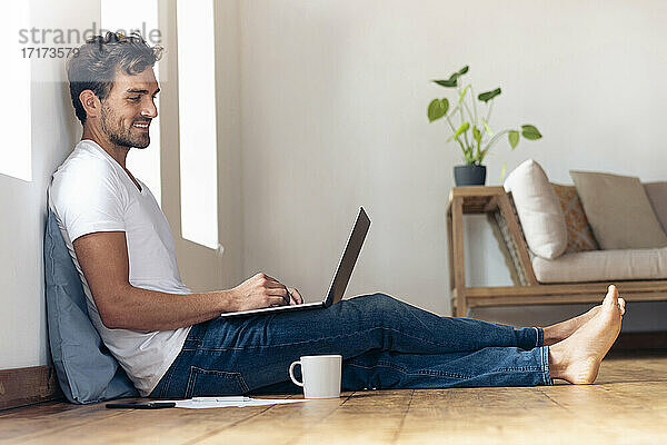 Lächelnder Mann  der einen Laptop benutzt  während er zu Hause auf dem Boden sitzt
