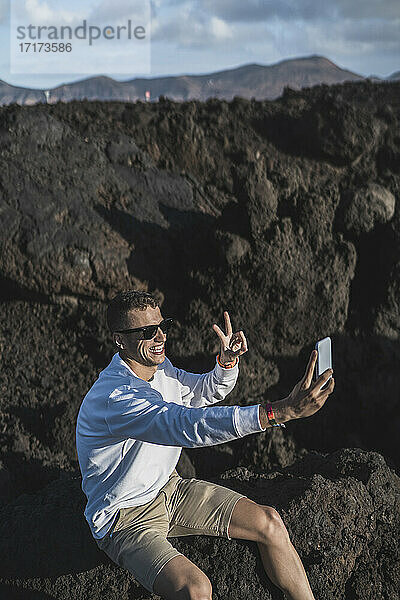 Männlicher Tourist  der ein Selfie macht  während er auf einem vulkanischen Felsen in Los Hervideros  Lanzarote  Spanien sitzt