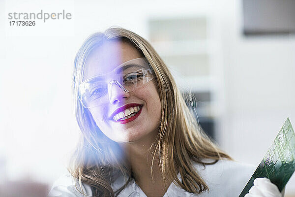 Lächelnde Technikerin mit Schutzbrille hält eine Leiterplatte im Labor