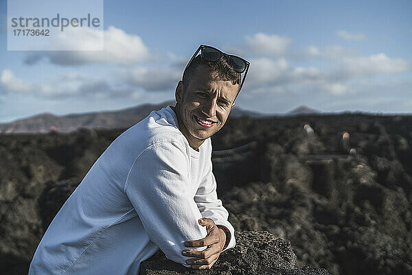 Lächelnder männlicher Tourist  der sich an einen Vulkanfelsen lehnt  an einem sonnigen Tag in Los Hervideros  Lanzarote  Spanien