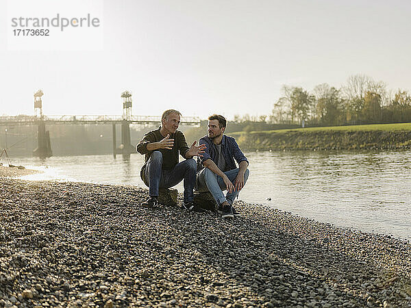 Father and son talking with each other while sitting on rock at riverbank during autumn