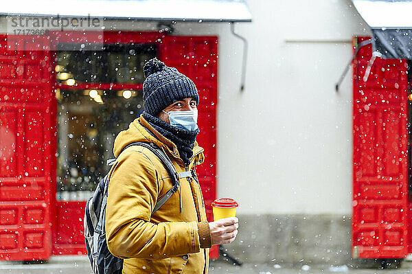 Man with coffee cup standing against store while snowing during pandemic