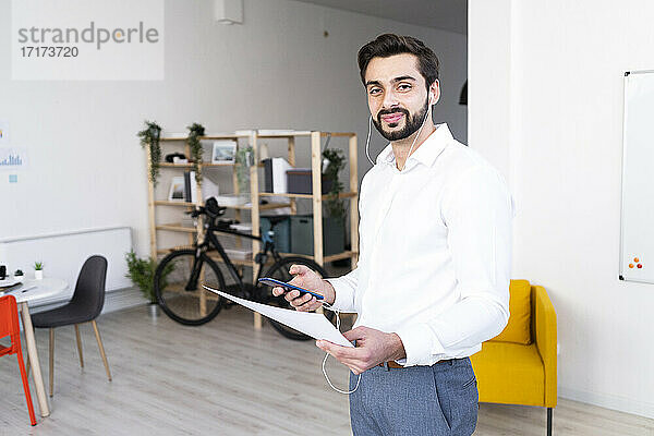 Lächelnde männliche Fachkraft mit Papierdokument  die ein Mobiltelefon bei der Arbeit am Arbeitsplatz hält