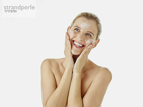 Smiling woman rubbing cream on face while standing against white background