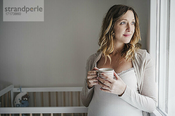 Nachdenkliche schwangere Frau  die eine Kaffeetasse hält  während sie zu Hause an der Krippe steht