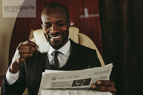 Glücklicher männlicher Unternehmer mit Kaffeetasse beim Lesen der Zeitung im Privatjet