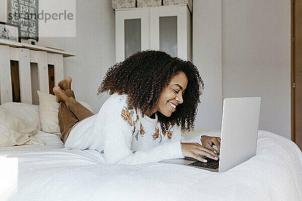 Junge Frau lächelt  während sie einen Laptop benutzt und zu Hause auf dem Bett liegt