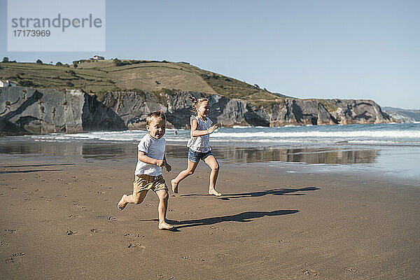 Playful brother and sister running at beach against hill during sunny day
