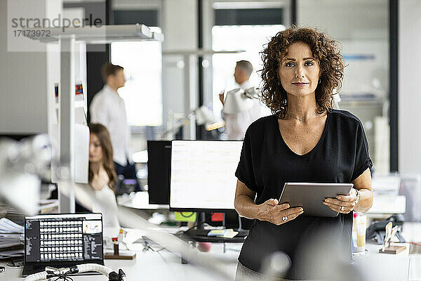 Unternehmerin hält digitales Tablet  während sie mit einem Kollegen im Hintergrund in einem Großraumbüro steht