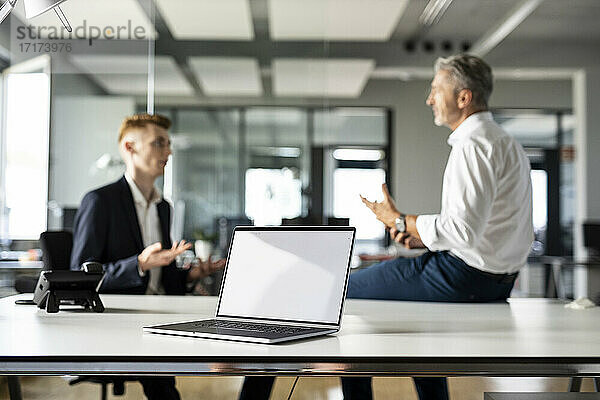 Laptop auf einem Tisch mit Geschäftsleuten  die im Hintergrund in einem Großraumbüro diskutieren