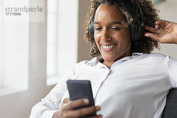 Lächelnde Frau  die Musik hört  während sie ein Mobiltelefon benutzt und zu Hause sitzt