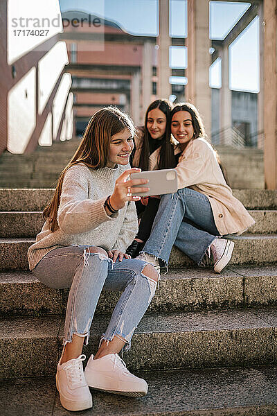 Lächelndes Teenager-Mädchen  das ein Selfie mit Freunden macht  während es auf einer Treppe in der Stadt sitzt