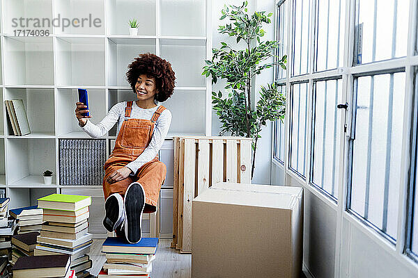 Lächelnde Afro-Frau  die ein Selfie vor einem Bücherregal macht  während sie in eine neue Loft-Wohnung zieht
