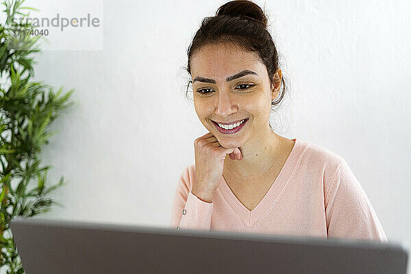 Smiling woman with hand on chin working on laptop while sitting at home