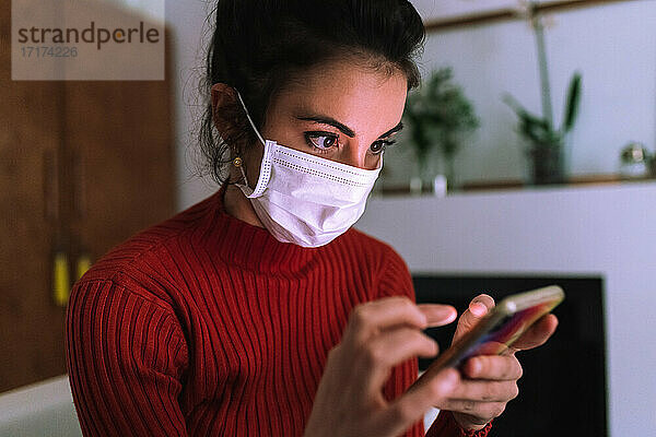 Junge Frau mit Coronavirus-Gesichtsmaske  die auf ihr Telefon schaut