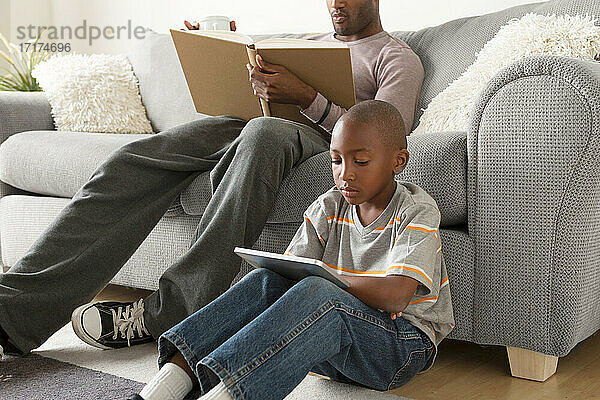 Junge sitzt auf dem Boden und benutzt ein digitales Tablet  Vater liest ein Buch