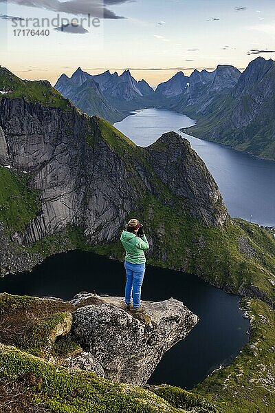 Abendstimmung  junge Wanderin steht auf Felsvorsprung und fotografiert die Aussicht vom Reinebringen  Reinebriggen  Hamnoy  Reine und den Reinefjord mit Bergen  Moskenes  Moskenesöy  Lofoten  Norwegen  Europa