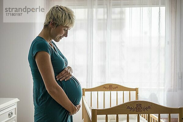Schwangere Frau hält ihren Bauch durch Babybett neben dem Fenster