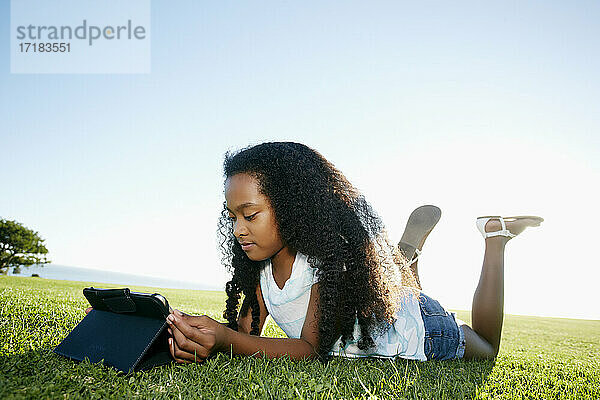 Neun Jahre altes  gemischtrassiges Mädchen liegt im Gras und schaut auf ein digitales Tablet.