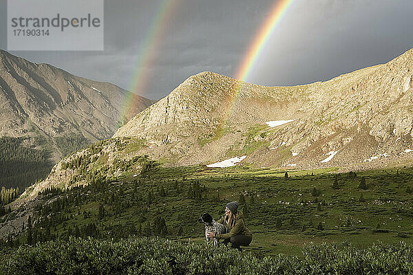 Frau streichelt Hund  während doppelter Regenbogen und Berge im Hintergrund
