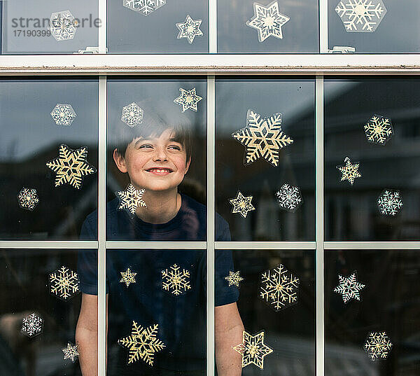 Fröhlicher Junge  der durch ein mit Schneeflocken bedecktes Fenster hinausschaut.