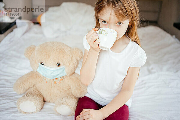 Mädchen trinkt Tee mit einem Teddybär in einer medizinischen Maske auf einem weißen Bett