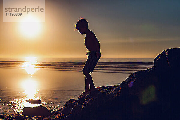 Silhouette eines Jungen  der bei Sonnenuntergang die Felsen am Strand hinunterläuft.