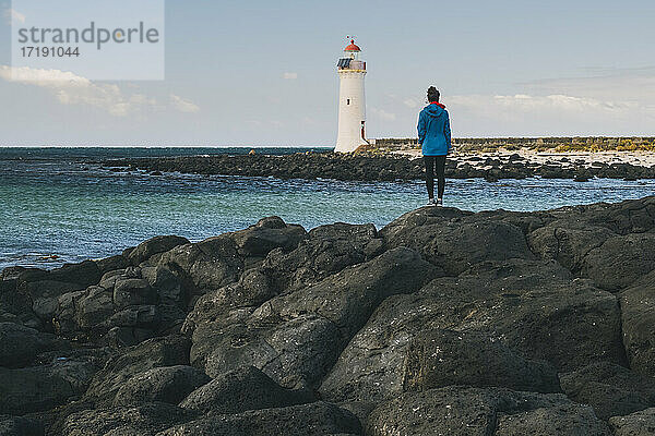 Junge Frau mit Blick auf einen Leuchtturm in der Ferne  der auf der felsigen Küste steht  Port Fairy  Australien.