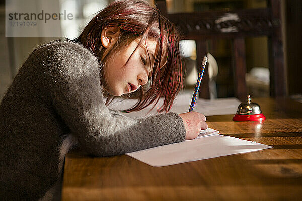 Konzentriertes kleines Mädchen lehnt sich über Papier und schreibt mit Bleistift am Tisch