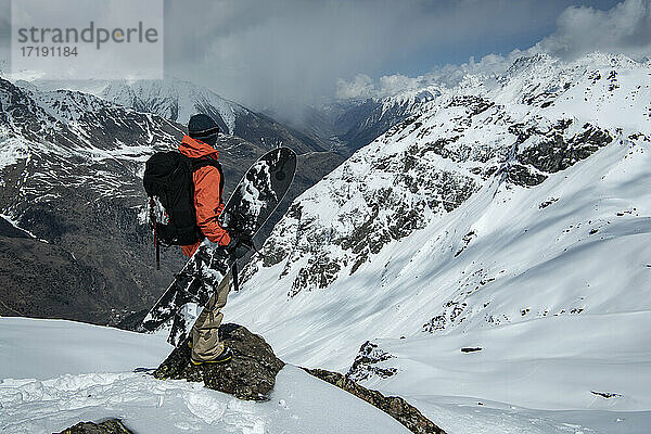 Mann mit Snowboard steht auf einem Felsen an einem schneebedeckten Berg gegen einen bewölkten Himmel