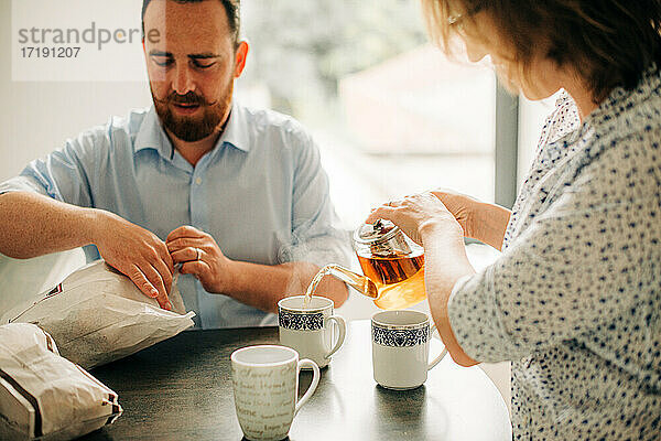 Ehepaar beim Frühstück mit Tee. Mann öffnet Tüten mit frischem Gebäck
