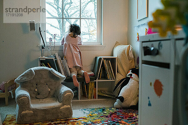 Junges Mädchen in unordentlichem Zimmer mit Spielzeug schaut aus dem Fenster