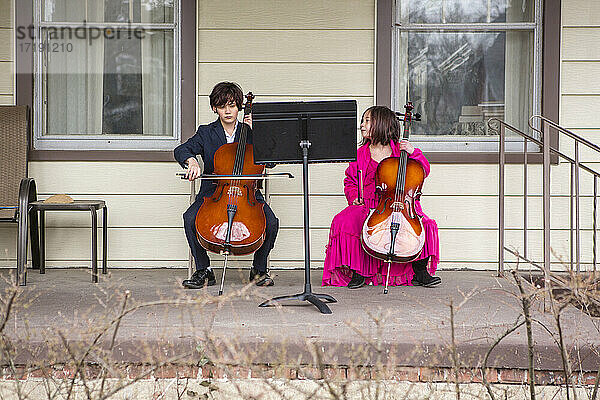 Ein Junge sitzt auf der Veranda und spielt Cello im Anzug  während seine Schwester zusieht
