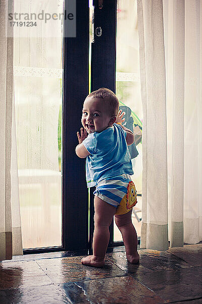 Kleiner barfüßiger Junge hält sich an einer großen Glastür fest.