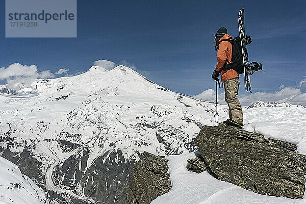 Mann mit Skistock und Snowboard steht auf einem Felsen vor einem schneebedeckten Berg gegen den Himmel