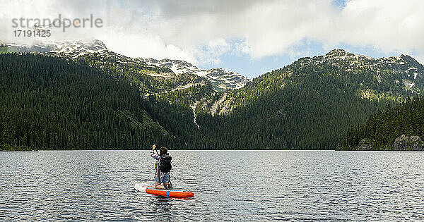 Rückenansicht einer Frau  die auf einem Paddleboard kniet und auf dem kräuselnden Wasser eines Sees in Whistler  Kanada  in Richtung der grünen Berge paddelt