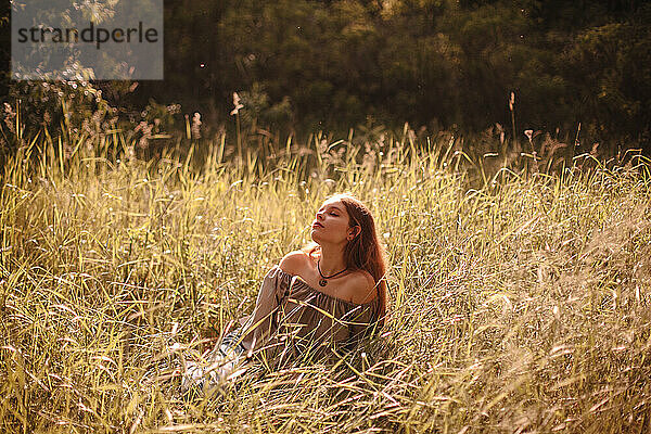 Friedliche junge Frau  die im Sommer auf einem Feld im Gras sitzt